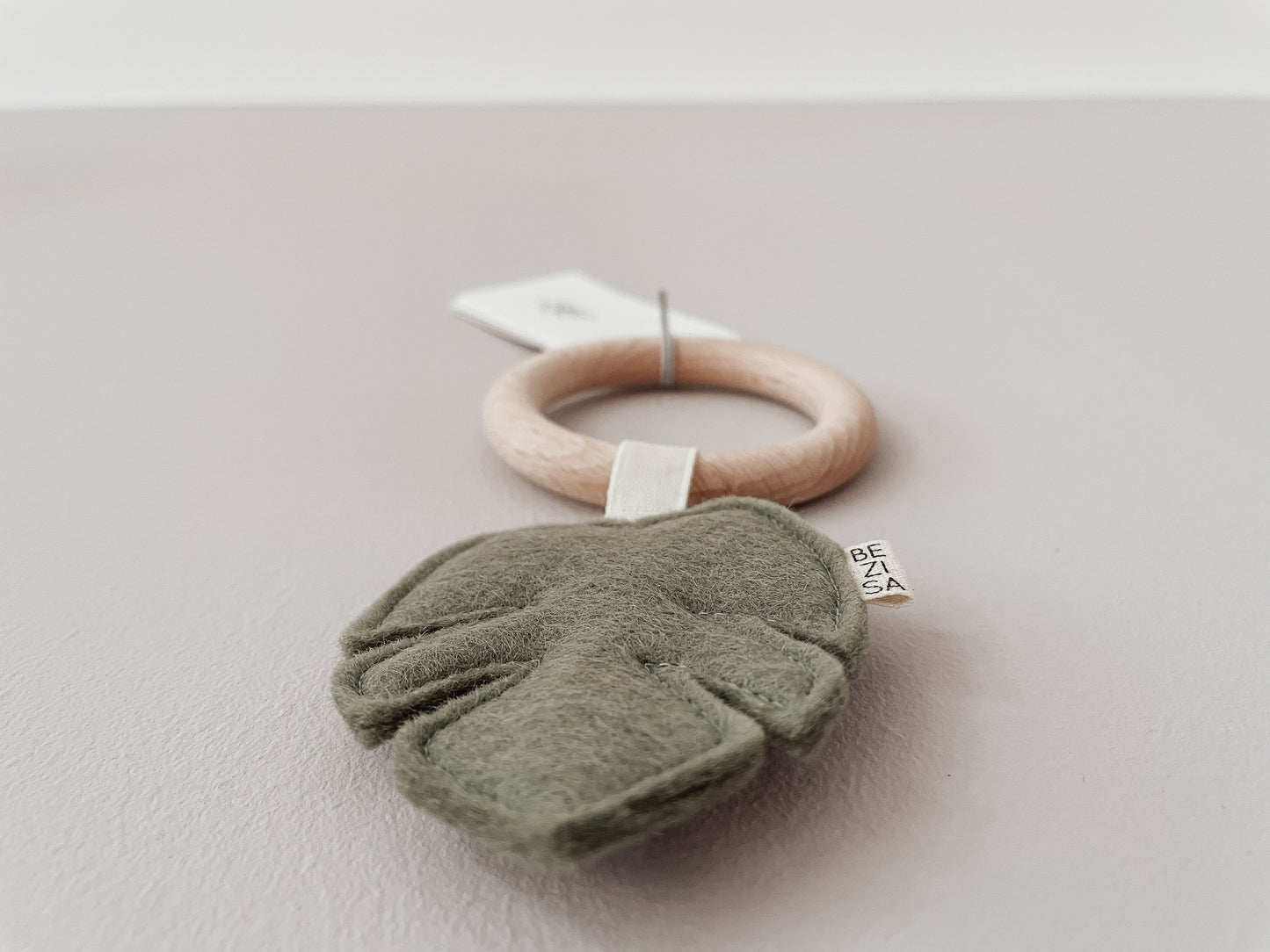 Baby teether from Bezisa, monstera leaf in pistachio green felt with a beech wood ring. Photographed on a plain neutral background.