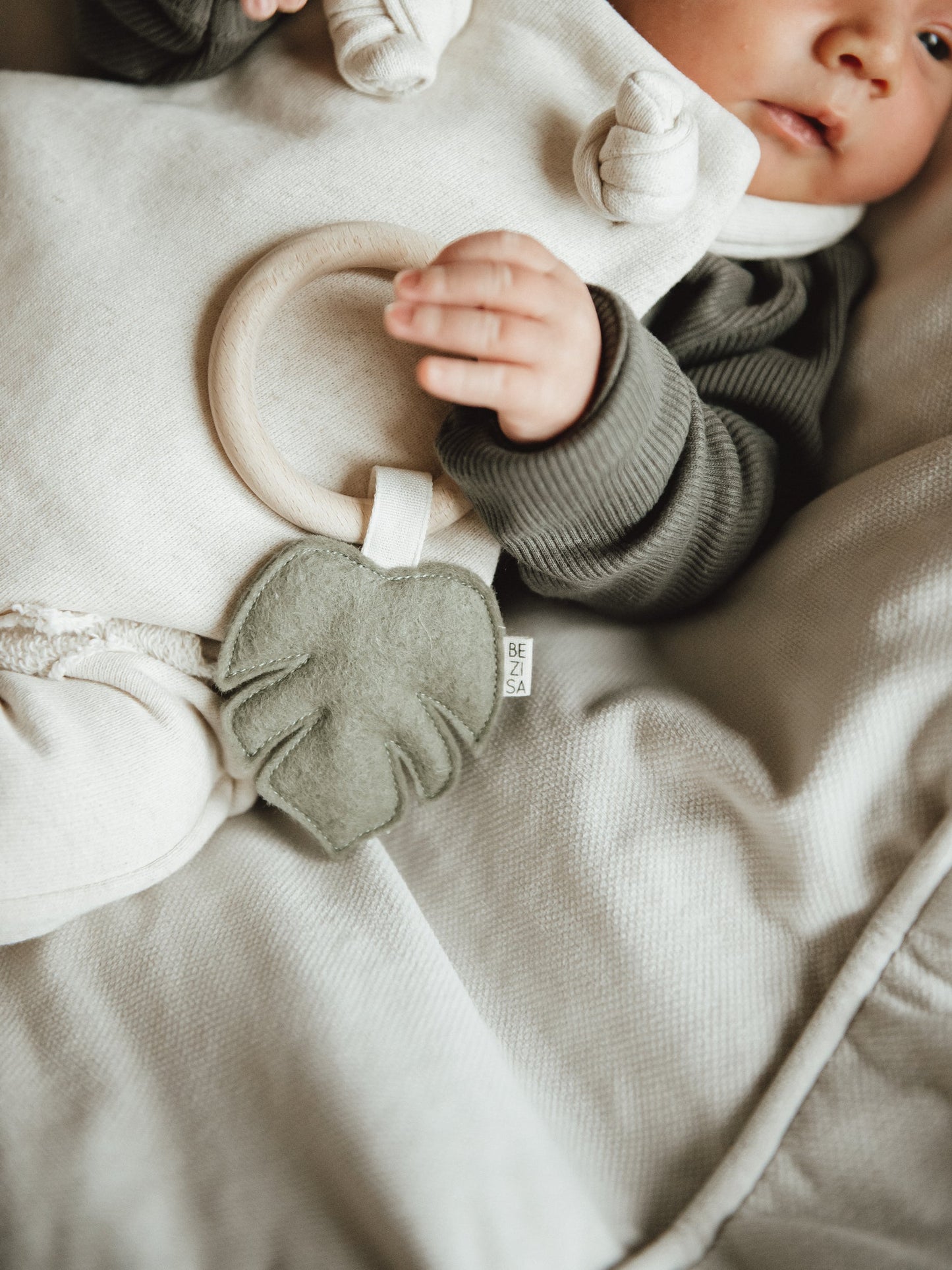 Baby teether from Bezisa, monstera leaf in pistachio green felt with a beech wood ring. Baby laying down is holding the teether.