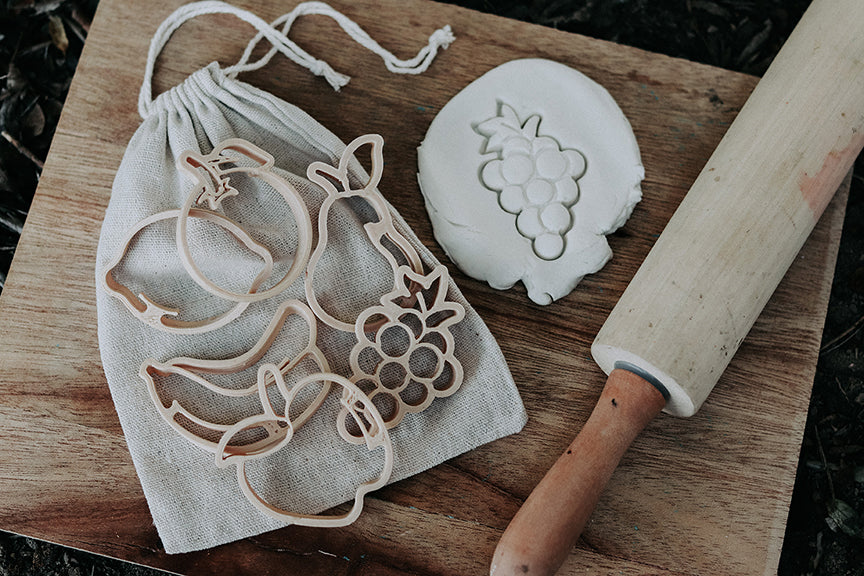 Kinfolk Pantry set of 6 mini fruit eco cutters photographed on a cotton drawstring bag on some wood with a rolling pin and the grapes cutter already used to create the shape in the white playdough. Cutter set includes bananas, orange, apple, lemon, grapes and pear.
