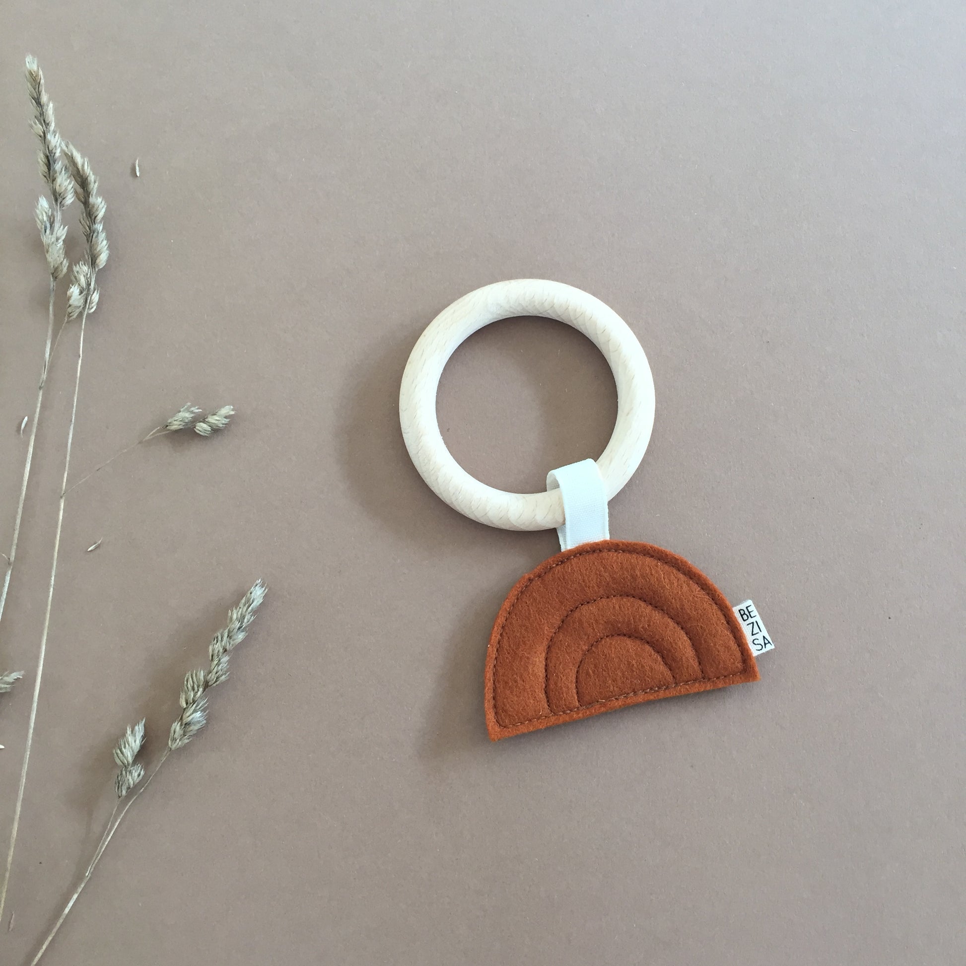 Bezisa Sunset teether in rust colour with wooden teething ring photographed on a plain neutral background with some dried flowers.