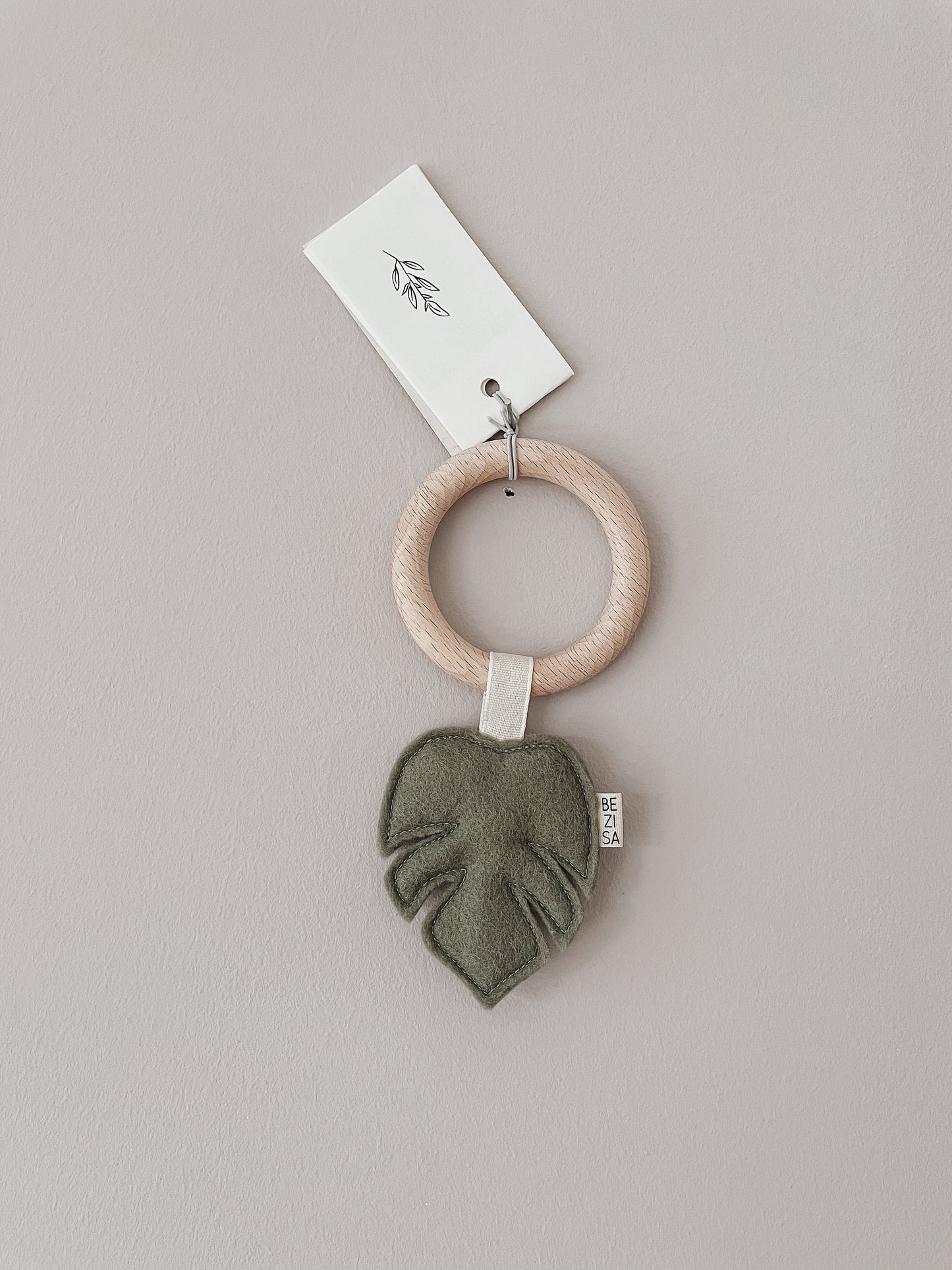 Baby teether from Bezisa, monstera leaf in pistachio green felt with a beech wood ring. Photographed on a plain neutral background.