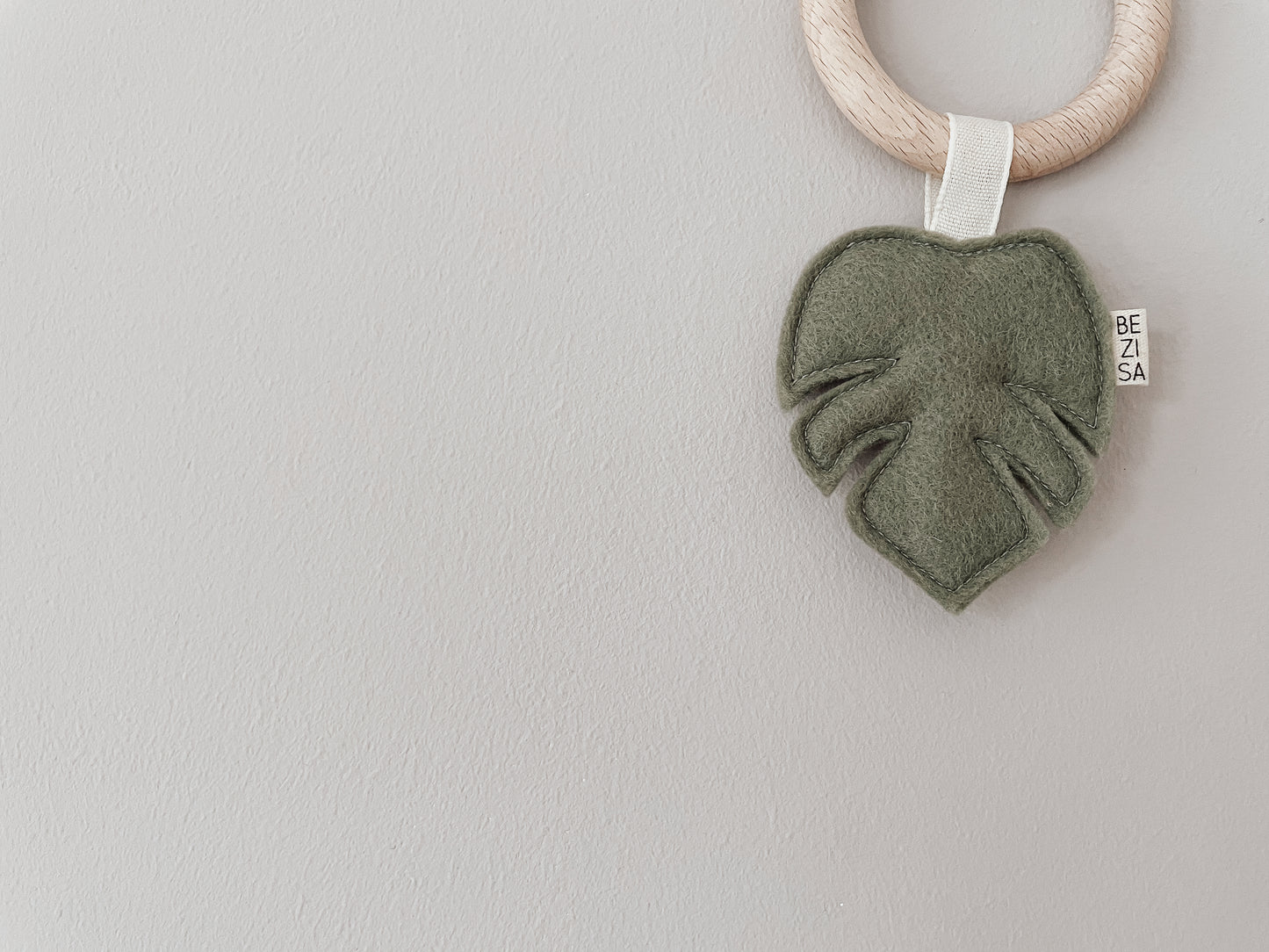 Baby teether from Bezisa, monstera leaf in pistachio green felt with a beech wood ring. Photographed on a plain neutral background.
