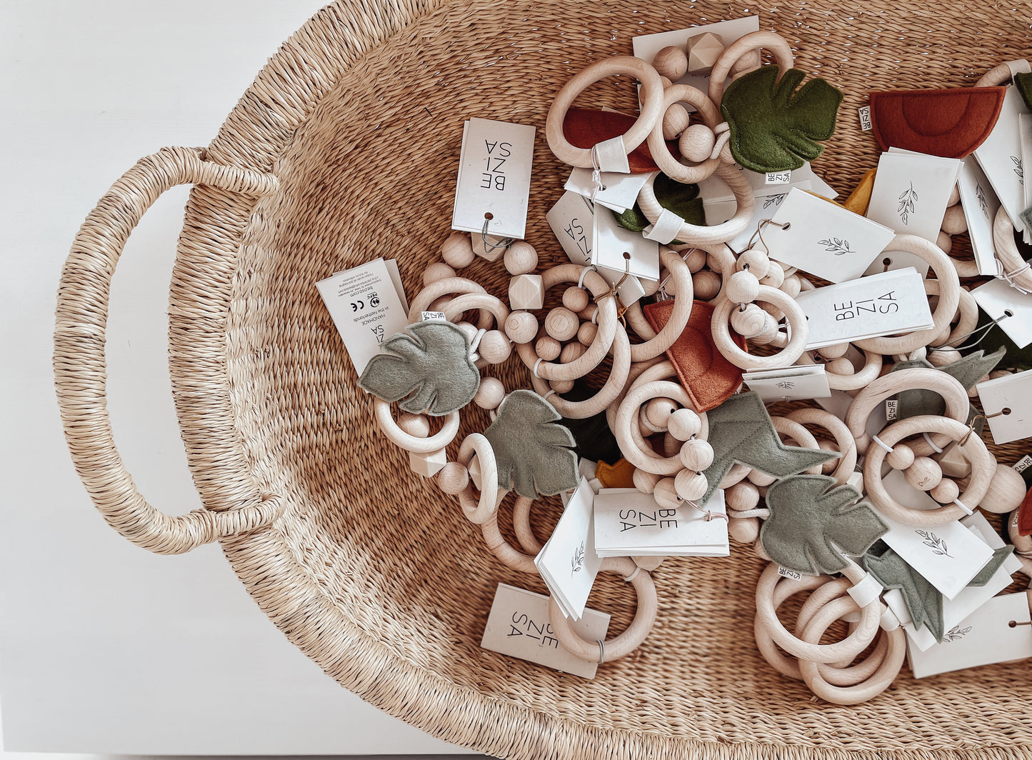 A collection of baby teethers and wooden rattles from Bezisa. A mix of monstera leaf in pistachio green felt with a beech wood ring teether, rust colour sunset rainbow teether, all in a wicker basket.