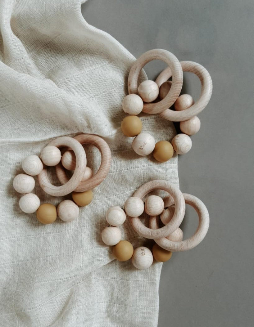 Bezisa wooden baby rattle with a mix of round wooden beads and camel coloured 100% silicone beads, plus two wooden rings. Three rattles are photographed on a neutral muslin.