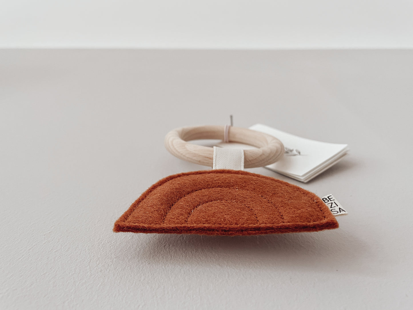 Bezisa Sunset teether in rust colour with wooden teething ring, photographed on a plain neutral background.