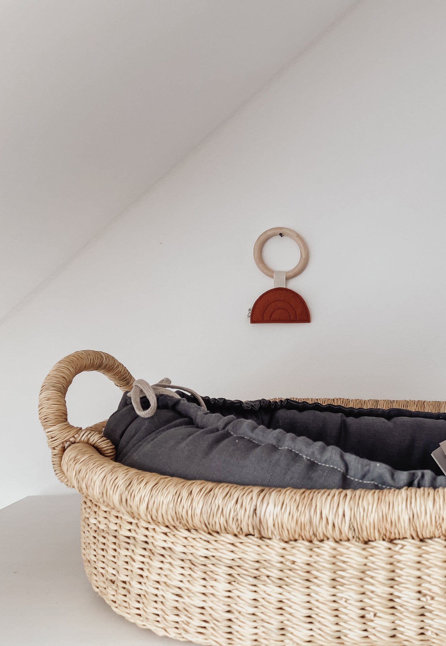 Bezisa Sunset teether in rust colour with wooden teething ring, hanging on a plain white wall. Baby changing basket is in the foreground.