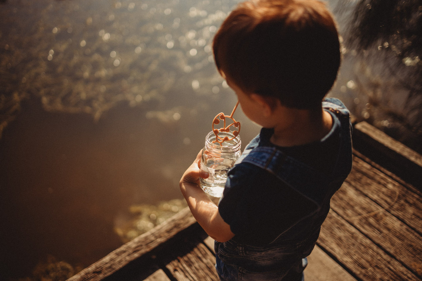 A boy in denim dungarees is dipping the ladybird shaped eco bubble wand from Kinfolk Pantry into a jar of magic potion ready to blow bubbles! The boy is standing on a boardwalk in an outdoor forest setting with a lake.