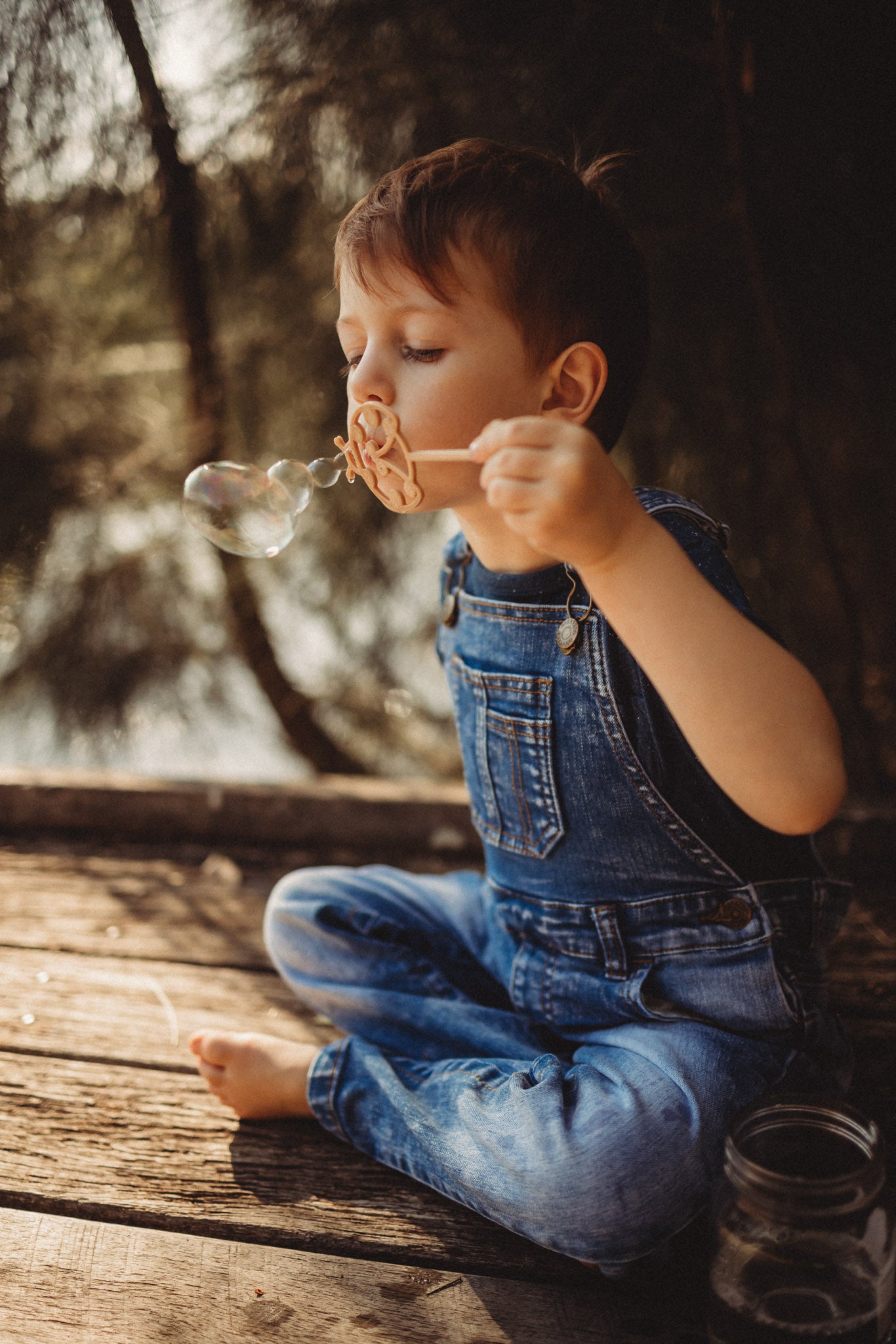 A boy in denim dungarees is blowing bubbles through the ladybird shaped eco bubble wand from Kinfolk Pantry. The boy is sitting on a boardwalk in an outdoor forest setting.