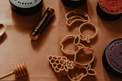 Kinfolk Pantry set of 6 mini fruit eco cutters surrounded by tins of playdough ready for sensory learning and play. Cutter set includes bananas, orange, apple, lemon, grapes and pear.