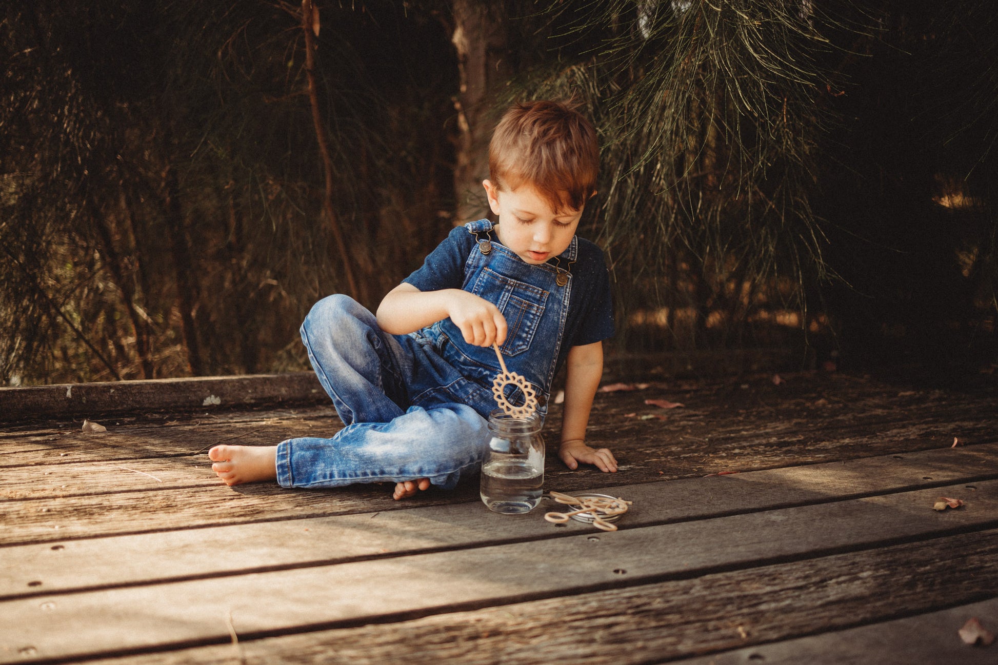 A boy in blue dungarees is sitting on a boardwalk dipping the sunflower shaped eco bubble wand from Kinfolk Pantry into some magic potion to make bubbles with!