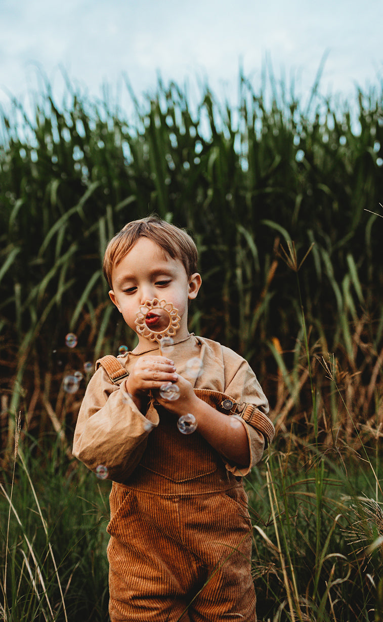 A boy in rust colour dungarees is holding and blowing bubbles through the sunflower shaped eco bubble wand from Kinfolk Pantry - there are a few bubble that haven't yet been popped!