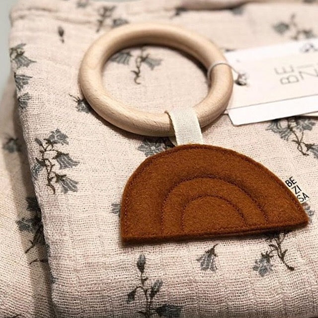 Bezisa Sunset teether in rust colour with wooden teething ring laying on a neutral floral muslin swaddle.