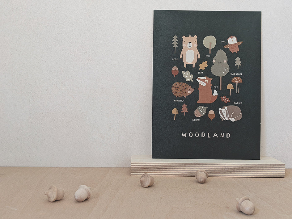 Nursery wall art print with dark moss green background with a muted simple design of animal illustrations and things you'd find on a nature walk. Trees, leaves, acorns, toadstools, bear, owl, fox, hedgehog, badger. Little wooden acorn decorations in the foreground of the photo.