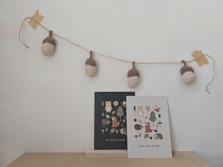 Woodland animals and nature illustrations, A4 art prints for the nursery or playroom. Two options, neutral pebble background or dark moss green. Prints are sitting in a wooden display stand against a wall where a garland of knitted acorns is hung up on the wall. Simple, Scandi style, hygge living.