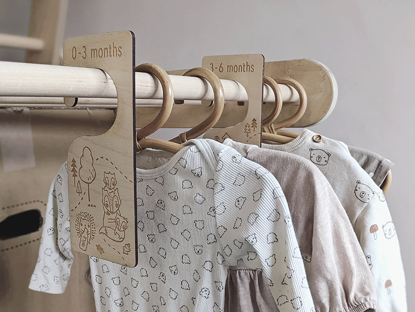 Neutral baby clothes are hanging on a wooden rail with baby clothing organisers dividing the ages from newborn to 12 months. These wooden hangers are perfect for keeping baby clothes organised.