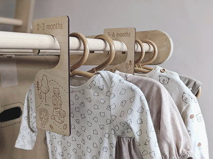 Neutral baby clothes are hanging on a wooden rail with baby clothing organisers dividing the ages from newborn to 12 months. These wooden hangers are perfect for keeping baby clothes organised.