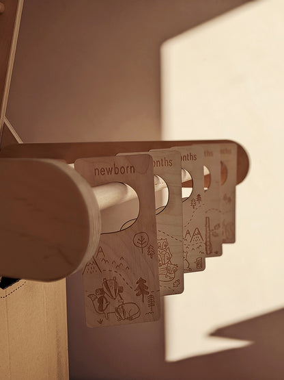 Lots of sunlight shining onto wooden baby clothing divider hangers from Paper & Bean. Each hanger has a woodland scene laser cut into the plywood. There are five dividers from newborn, 0-3 months, 3-6 months, 6-9 months, 9-12 months.